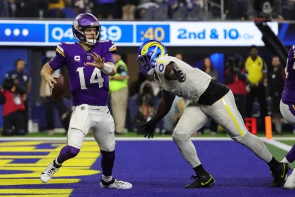 NFL Playoffs & NFL Offseason: Ein Footballspieler in violetter Uniform und mit Helmnummer 14 bereitet sich darauf vor, den Ball zu werfen, während er von einem gegnerischen Spieler in weißer Uniform und mit blau-gelbem Helm (Nummer 90) verfolgt wird. Die spannende Action spielt sich während der NFL-Playoffs auf einem blauen Feld ab, im Hintergrund ist eine Markierung für den 2. und 10. Versuch zu sehen. Diese Beschreibung wurde mit der FootballR KI automatisch generiert.