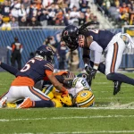 Jordan Love verletzt. Zwei Spieler der Chicago Bears tackeln während eines spannenden Footballspiels einen Spieler der Green Bay Packers, bei dem es sich um Christian Watson handeln könnte. Während der Spieler in Grün mit dem Ball im Griff auf dem Rasen liegt, füllen sich die Tribünen mit gespannten Zuschauern und die Offiziellen beobachten aufmerksam. Die Bears tragen marineblaue und orangefarbene Uniformen, die Packers tragen grün und gelb. Diese Beschreibung wurde mit der FootballR KI automatisch generiert.