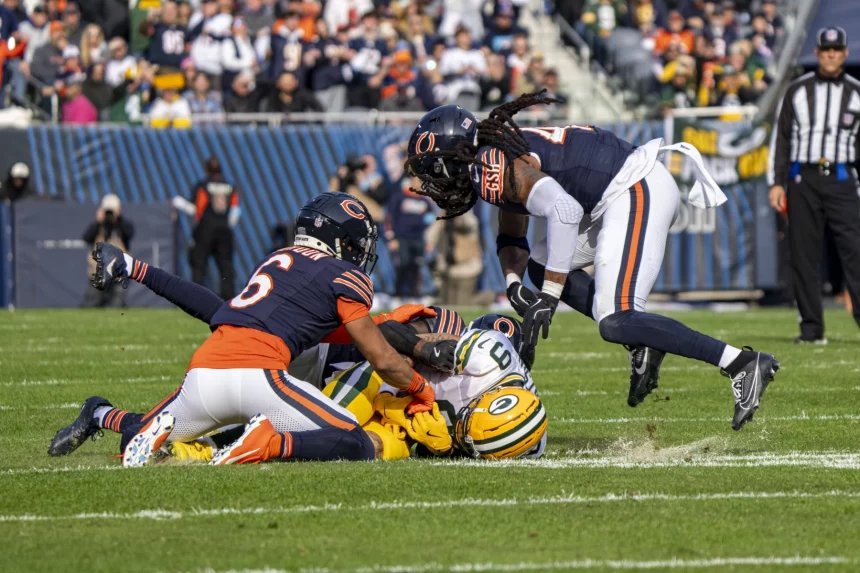 Jordan Love verletzt. Zwei Spieler der Chicago Bears tackeln während eines spannenden Footballspiels einen Spieler der Green Bay Packers, bei dem es sich um Christian Watson handeln könnte. Während der Spieler in Grün mit dem Ball im Griff auf dem Rasen liegt, füllen sich die Tribünen mit gespannten Zuschauern und die Offiziellen beobachten aufmerksam. Die Bears tragen marineblaue und orangefarbene Uniformen, die Packers tragen grün und gelb. Diese Beschreibung wurde mit der FootballR KI automatisch generiert.