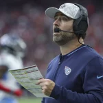 Ein Footballtrainer in marineblauer Jacke, Tennessee Titans Kappe und Headset hält ein Spielblatt in der Hand und blickt nach oben. Im Hintergrund ist ein Footballfeld mit verschwommenen Figuren zu sehen, darunter ein Spieler in weiß-blauer Uniform. Die Szene lässt auf ein laufendes Spiel mit Zuschauern schließen. Diese Beschreibung wurde mit der FootballR KI automatisch generiert.