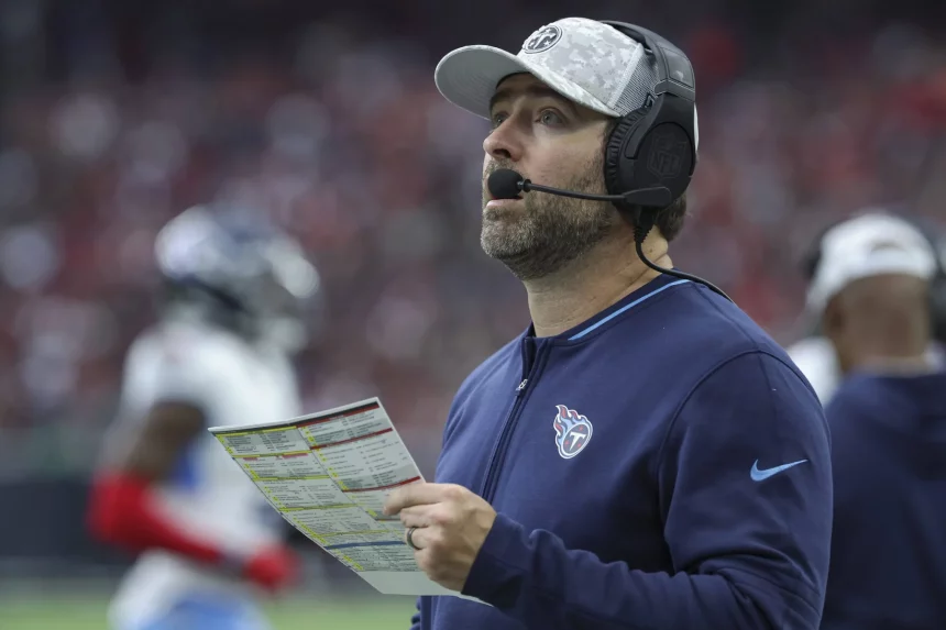 Ein Footballtrainer in marineblauer Jacke, Tennessee Titans Kappe und Headset hält ein Spielblatt in der Hand und blickt nach oben. Im Hintergrund ist ein Footballfeld mit verschwommenen Figuren zu sehen, darunter ein Spieler in weiß-blauer Uniform. Die Szene lässt auf ein laufendes Spiel mit Zuschauern schließen. Diese Beschreibung wurde mit der FootballR KI automatisch generiert.