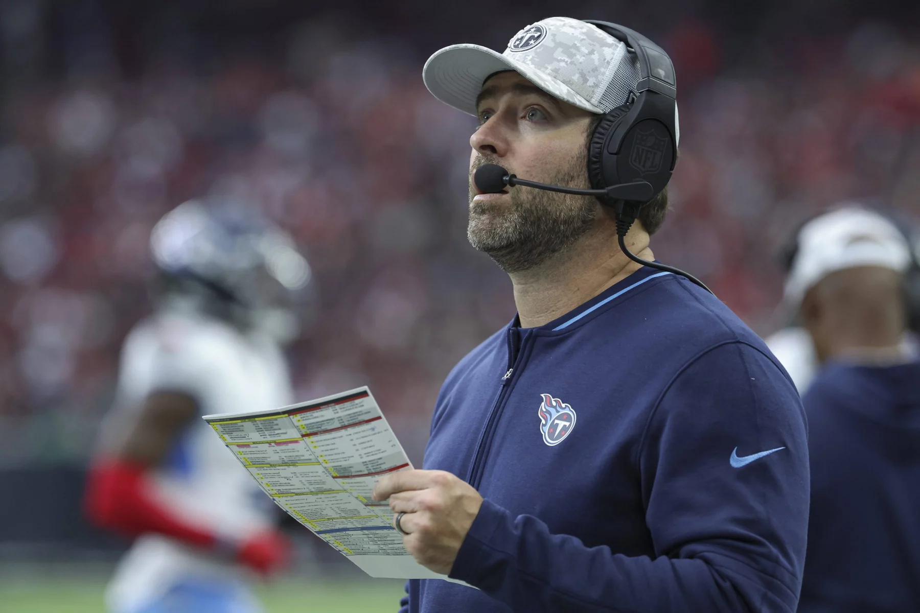 Ein Footballtrainer in marineblauer Jacke, Tennessee Titans Kappe und Headset hält ein Spielblatt in der Hand und blickt nach oben. Im Hintergrund ist ein Footballfeld mit verschwommenen Figuren zu sehen, darunter ein Spieler in weiß-blauer Uniform. Die Szene lässt auf ein laufendes Spiel mit Zuschauern schließen. Diese Beschreibung wurde mit der FootballR KI automatisch generiert.