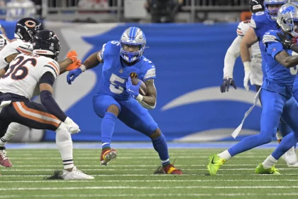 David Montgomery, in blauer Uniform und Helm, läuft mit dem Ball und weicht Verteidigern in schwarz-orangefarbenen Uniformen gekonnt aus. Das Spiel findet auf einem grünen Feld mit einer verschwommenen Menschenmenge statt, während seine Teamkollegen in der Nähe entscheidende Unterstützung leisten. Diese Beschreibung wurde mit der FootballR KI automatisch generiert.