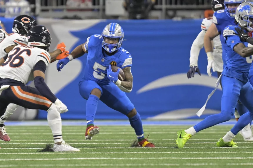 David Montgomery, in blauer Uniform und Helm, läuft mit dem Ball und weicht Verteidigern in schwarz-orangefarbenen Uniformen gekonnt aus. Das Spiel findet auf einem grünen Feld mit einer verschwommenen Menschenmenge statt, während seine Teamkollegen in der Nähe entscheidende Unterstützung leisten. Diese Beschreibung wurde mit der FootballR KI automatisch generiert.