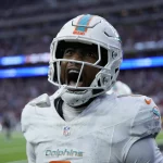 Jonnu Smith in der weißen Uniform der Miami Dolphins schreit leidenschaftlich auf dem Feld und verkörpert Jonnu Smiths Intensität. Der Hintergrund zeigt ein verschwommenes Stadion mit hellen Lichtern und einer jubelnden Menge, das die Energie und Aufregung des Spiels einfängt. Diese Beschreibung wurde mit der FootballR KI automatisch generiert.