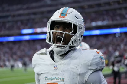 Jonnu Smith in der weißen Uniform der Miami Dolphins schreit leidenschaftlich auf dem Feld und verkörpert Jonnu Smiths Intensität. Der Hintergrund zeigt ein verschwommenes Stadion mit hellen Lichtern und einer jubelnden Menge, das die Energie und Aufregung des Spiels einfängt. Diese Beschreibung wurde mit der FootballR KI automatisch generiert.