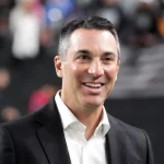 Ein Mann in einem schwarzen Blazer und einem weißen Hemd, das an Tom Telescos unverwechselbaren Stil erinnert, lächelt breit. Er hat kurzes, dunkles Haar und steht in einer verschwommenen, möglicherweise in einem Innenraum liegenden Szene mit Menschen im Hintergrund, was eine Aura dynamischen Umbruchs suggeriert. Diese Beschreibung wurde mit der FootballR KI automatisch generiert.