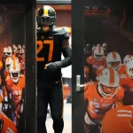 James Pearce Jr. trägt eine schwarz-orangefarbene Uniform mit der Nummer 27 und tritt durch eine Tür. In der Tür ist ein Foto seines Teams in orange-weißen Uniformen mit sichtbaren Nummern und Helmen zu sehen, das durch einen Feldtunnel geht. Die Szene lässt auf ein Spiel oder eine Trainingsumgebung schließen. Diese Beschreibung wurde mit der FootballR KI automatisch generiert.