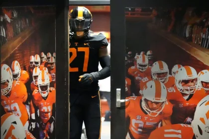 James Pearce Jr. trägt eine schwarz-orangefarbene Uniform mit der Nummer 27 und tritt durch eine Tür. In der Tür ist ein Foto seines Teams in orange-weißen Uniformen mit sichtbaren Nummern und Helmen zu sehen, das durch einen Feldtunnel geht. Die Szene lässt auf ein Spiel oder eine Trainingsumgebung schließen. Diese Beschreibung wurde mit der FootballR KI automatisch generiert.