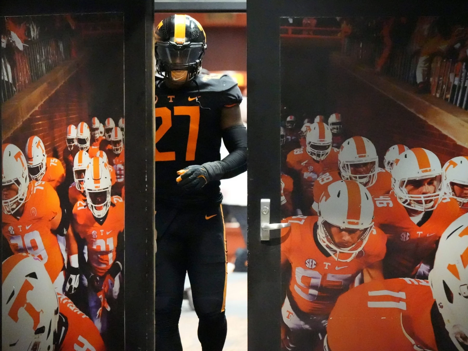 James Pearce Jr. trägt eine schwarz-orangefarbene Uniform mit der Nummer 27 und tritt durch eine Tür. In der Tür ist ein Foto seines Teams in orange-weißen Uniformen mit sichtbaren Nummern und Helmen zu sehen, das durch einen Feldtunnel geht. Die Szene lässt auf ein Spiel oder eine Trainingsumgebung schließen. Diese Beschreibung wurde mit der FootballR KI automatisch generiert.