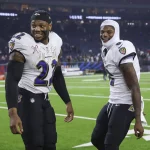 NFL Playoffs: Zwei Footballspieler in weißen Trikots und dunklen Hosen der Baltimore Ravens. Derrick Henry trägt eine schwarze Kappe und das Trikot mit der Nummer 22, während sein Teamkollege Lamar Jackson eine weiße Kapuze und das Trikot mit der Nummer 8 trägt. Im Hintergrund sorgen verschwommene Zuschauer und Sportgeräte für die Atmosphäre. Diese Beschreibung wurde mit der FootballR KI automatisch generiert.