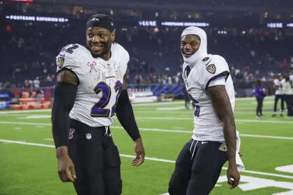 NFL Playoffs: Zwei Footballspieler in weißen Trikots und dunklen Hosen der Baltimore Ravens. Derrick Henry trägt eine schwarze Kappe und das Trikot mit der Nummer 22, während sein Teamkollege Lamar Jackson eine weiße Kapuze und das Trikot mit der Nummer 8 trägt. Im Hintergrund sorgen verschwommene Zuschauer und Sportgeräte für die Atmosphäre. Diese Beschreibung wurde mit der FootballR KI automatisch generiert.