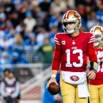 Ein Footballspieler, der an Brock Purdy erinnert, in einer rot-goldenen Uniform, Nummer 13, hält einen Football, während er über das Feld geht. Er trägt einen metallisch-goldenen Helm mit einem roten Streifen. Ein weiterer Spieler ist im Hintergrund zu sehen, während Zuschauer in Blau und Weiß die Tribünen füllen. Diese Beschreibung wurde mit der FootballR KI automatisch generiert.