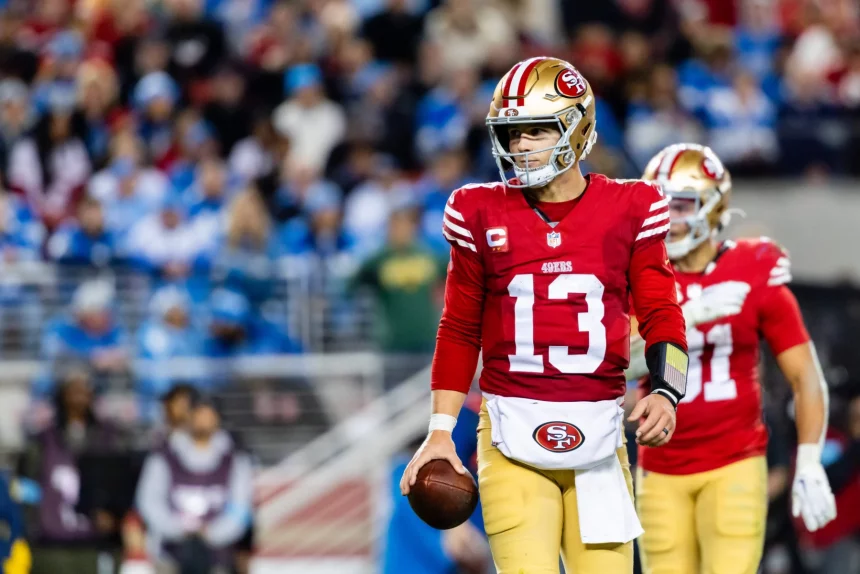 Ein Footballspieler, der an Brock Purdy erinnert, in einer rot-goldenen Uniform, Nummer 13, hält einen Football, während er über das Feld geht. Er trägt einen metallisch-goldenen Helm mit einem roten Streifen. Ein weiterer Spieler ist im Hintergrund zu sehen, während Zuschauer in Blau und Weiß die Tribünen füllen. Diese Beschreibung wurde mit der FootballR KI automatisch generiert.