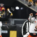 Joe Burrow in weißer Uniform wirft einen Pass, während ein Spieler in Schwarz seine Hände zum Blocken hebt. Das Stadion summt, im Hintergrund sind verschwommene Zuschauer zu sehen. Der konzentrierte Gesichtsausdruck des Quarterbacks fängt die Intensität ein, während beide Spieler mitten im Spiel sind. Diese Beschreibung wurde mit der FootballR KI automatisch generiert.