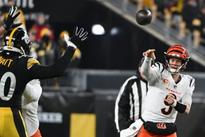 Joe Burrow in weißer Uniform wirft einen Pass, während ein Spieler in Schwarz seine Hände zum Blocken hebt. Das Stadion summt, im Hintergrund sind verschwommene Zuschauer zu sehen. Der konzentrierte Gesichtsausdruck des Quarterbacks fängt die Intensität ein, während beide Spieler mitten im Spiel sind. Diese Beschreibung wurde mit der FootballR KI automatisch generiert.