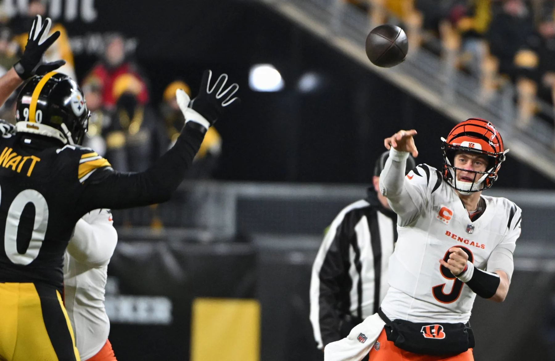 Joe Burrow in weißer Uniform wirft einen Pass, während ein Spieler in Schwarz seine Hände zum Blocken hebt. Das Stadion summt, im Hintergrund sind verschwommene Zuschauer zu sehen. Der konzentrierte Gesichtsausdruck des Quarterbacks fängt die Intensität ein, während beide Spieler mitten im Spiel sind. Diese Beschreibung wurde mit der FootballR KI automatisch generiert.