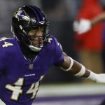 Marlon Humphrey, ein Ravens-Star in einer lila Baltimore Ravens-Uniform, stürmt entschlossen voran. Er trägt das Trikot mit der Nummer 44 und einen Helm mit Visier und seine Arme sind zum Blocken oder Tacklen bereit. Der Hintergrund zeigt einen Teil des Felds und einen weiteren Spieler und spiegelt so die intensive Spielaktion wider. Diese Beschreibung wurde mit der FootballR KI automatisch generiert.