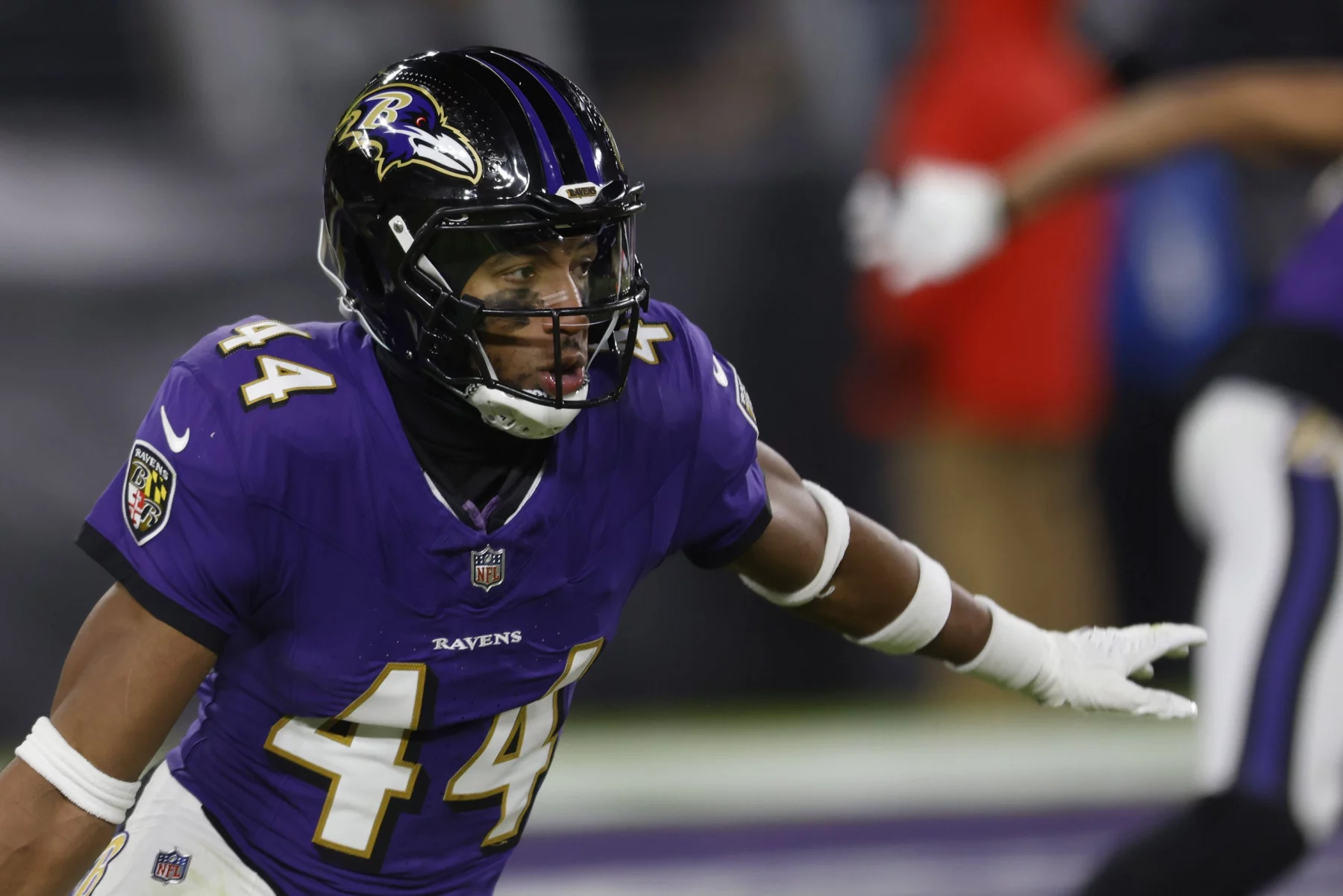 Marlon Humphrey, ein Ravens-Star in einer lila Baltimore Ravens-Uniform, stürmt entschlossen voran. Er trägt das Trikot mit der Nummer 44 und einen Helm mit Visier und seine Arme sind zum Blocken oder Tacklen bereit. Der Hintergrund zeigt einen Teil des Felds und einen weiteren Spieler und spiegelt so die intensive Spielaktion wider. Diese Beschreibung wurde mit der FootballR KI automatisch generiert.