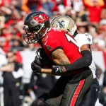 Ein Footballspieler im roten Trikot der Tampa Bay Buccaneers mit der Nummer 13 fängt einen Pass, während er von einem Spieler im weißen Trikot angegriffen wird. Sie sind auf dem Spielfeld, im Hintergrund ist eine verschwommene Menschenmenge zu sehen, die die Action und Intensität dieses spannenden Spiels einfängt. Diese Beschreibung wurde mit der FootballR KI automatisch generiert.