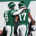 Zwei Footballspieler in grünen Uniformen der New York Jets feiern auf dem Spielfeld. Der Spieler links, Aaron Rodgers, mit der Trikotnummer 8, hat seinen Arm um den Spieler rechts mit der Trikotnummer 17 gelegt. Beide tragen Helme und blicken von der Kamera weg auf einen großen weiß-grünen Hintergrund. Diese Beschreibung wurde mit der FootballR KI automatisch generiert.