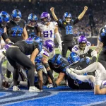 NFL Power Rankings - Footballspieler zweier Mannschaften in schwarz-blauen und violett-weißen Trikots drängen sich in der Nähe der Torlinie. Ein Spieler der Detroit Lions in Schwarz hält einen Football über die Linie, während andere ihn entweder feiern oder versuchen, ihn aufzuhalten. Die Menge im Hintergrund ist verschwommen. Diese Beschreibung wurde mit der FootballR KI automatisch generiert.