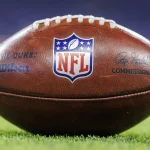 Wilson Football - Free Agency - Nahaufnahme eines NFL-Footballs, der auf einem Rasenfeld liegt. Der Ball zeigt in der Mitte das NFL-Logo, links das „THE DUKE“-Wilson-Abzeichen und rechts die Unterschrift des Commissioners. Obwohl das Design Aufmerksamkeit erregt, bleiben in Diskussionen zur Sicherheit Bedenken hinsichtlich Gehirnerschütterungen bestehen. Diese Beschreibung wurde mit der FootballR KI automatisch generiert.
