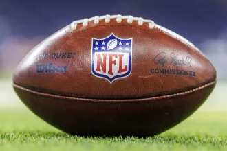 Wilson Football - Free Agency - Nahaufnahme eines NFL-Footballs, der auf einem Rasenfeld liegt. Der Ball zeigt in der Mitte das NFL-Logo, links das „THE DUKE“-Wilson-Abzeichen und rechts die Unterschrift des Commissioners. Obwohl das Design Aufmerksamkeit erregt, bleiben in Diskussionen zur Sicherheit Bedenken hinsichtlich Gehirnerschütterungen bestehen. Diese Beschreibung wurde mit der FootballR KI automatisch generiert.