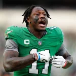 A.J. Brown in einem grünen Trikot mit der Nummer 11 und schwarzen Streifen unter den Augen gestikuliert energisch auf dem Feld. Er verkörpert die Intensität von A.J. Brown während dieses Playoff-Spiels. Mit grauen Handschuhen und einem Helm mit Logoaufklebern strahlt er vor dem verschwommenen Hintergrund eines spannenden Spiels. Diese Beschreibung wurde mit der FootballR KI automatisch generiert.