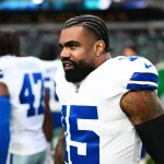Auf dem Feld steht Ezekiel Elliott in weiß-blauer Uniform mit der Nummer 15. Er hat geflochtene Haare und Tattoos auf den Armen. Im Hintergrund sind andere Spieler und ein Stadion voller lebhafter Zuschauer zu sehen, die das Spiel miterleben. Diese Beschreibung wurde mit der FootballR KI automatisch generiert.