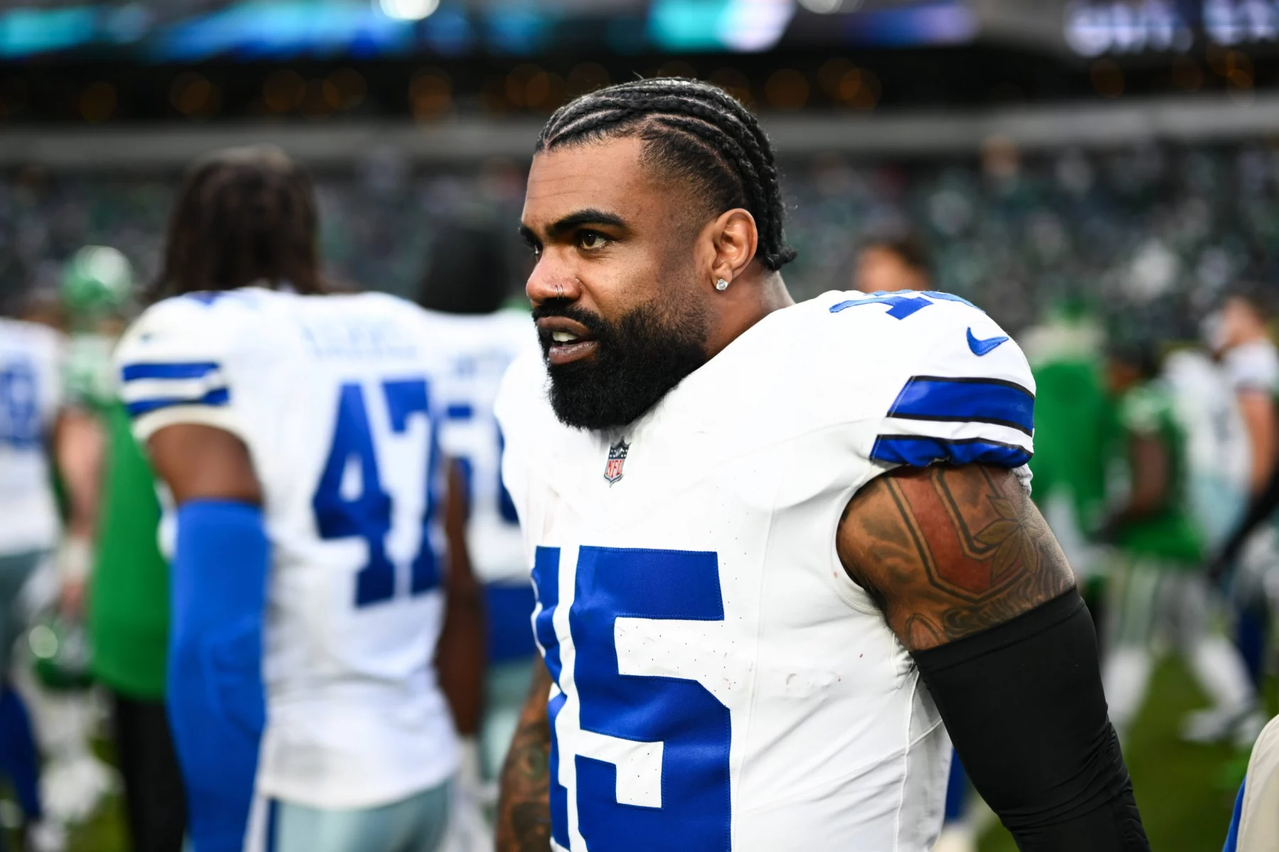 Auf dem Feld steht Ezekiel Elliott in weiß-blauer Uniform mit der Nummer 15. Er hat geflochtene Haare und Tattoos auf den Armen. Im Hintergrund sind andere Spieler und ein Stadion voller lebhafter Zuschauer zu sehen, die das Spiel miterleben. Diese Beschreibung wurde mit der FootballR KI automatisch generiert.