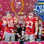 Footballspieler in rot-grauen Uniformen feiern einen Sieg auf der Bühne, während Konfetti herabregnet. Sie stehen neben Fans in Freizeitkleidung und sind von Auszeichnungen umgeben. Im Hintergrund sind Logos zu sehen, darunter „Goodyear“ und „Cotton Bowl“. Ein Spieler der Ohio State Buckeyes zeigt lächelnd nach oben. Diese Beschreibung wurde mit der FootballR KI automatisch generiert.