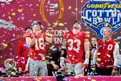 Footballspieler in rot-grauen Uniformen feiern einen Sieg auf der Bühne, während Konfetti herabregnet. Sie stehen neben Fans in Freizeitkleidung und sind von Auszeichnungen umgeben. Im Hintergrund sind Logos zu sehen, darunter „Goodyear“ und „Cotton Bowl“. Ein Spieler der Ohio State Buckeyes zeigt lächelnd nach oben. Diese Beschreibung wurde mit der FootballR KI automatisch generiert.