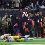 Ein Footballspieler in blau-weißer Uniform wird von einem anderen Spieler mit der Nummer 24, Derek Stingley Jr., zu Boden gerissen. Zuschauer und Fotografen fangen das intensive Spiel auf dem grünen Feld mit sichtbaren Yard-Linien ein. Diese Beschreibung wurde mit der FootballR KI automatisch generiert.