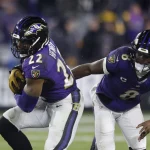 Zwei Footballspieler in lila Trikots stehen auf dem Feld. Der Spieler links, Derrick Henry, hält einen Football, trägt die Nummer 22 und rennt nach vorne. Der Spieler rechts, Lamar Jackson in Trikot Nummer 8, folgt ihm dicht auf den Fersen. Die verschwommene Menge füllt den Stadionhintergrund. Diese Beschreibung wurde mit der FootballR KI automatisch generiert.