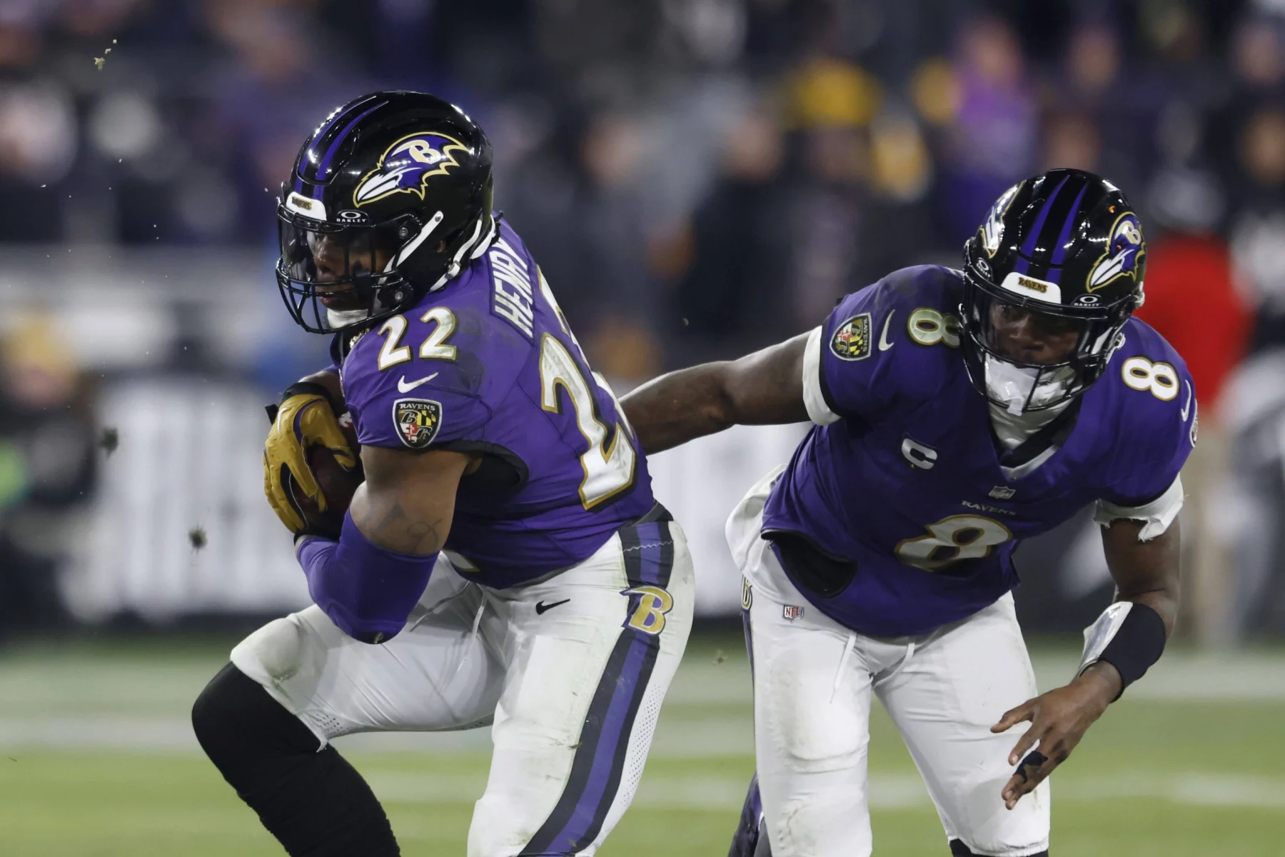 Zwei Footballspieler in lila Trikots stehen auf dem Feld. Der Spieler links, Derrick Henry, hält einen Football, trägt die Nummer 22 und rennt nach vorne. Der Spieler rechts, Lamar Jackson in Trikot Nummer 8, folgt ihm dicht auf den Fersen. Die verschwommene Menge füllt den Stadionhintergrund. Diese Beschreibung wurde mit der FootballR KI automatisch generiert.