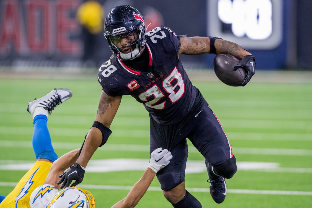 NFL-Star Joe Mixon, in einer schwarzen Uniform mit der Nummer 28, überholt gekonnt einen Gegner in einer weiß-gelben Uniform. Er rennt mit dem Ball über das leuchtend grüne Feld, weicht Tackles mühelos aus und demonstriert so die elektrisierende Intensität des Spiels. Diese Beschreibung wurde mit der FootballR KI automatisch generiert.