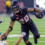 NFL-Star Joe Mixon, in einer schwarzen Uniform mit der Nummer 28, überholt gekonnt einen Gegner in einer weiß-gelben Uniform. Er rennt mit dem Ball über das leuchtend grüne Feld, weicht Tackles mühelos aus und demonstriert so die elektrisierende Intensität des Spiels. Diese Beschreibung wurde mit der FootballR KI automatisch generiert.