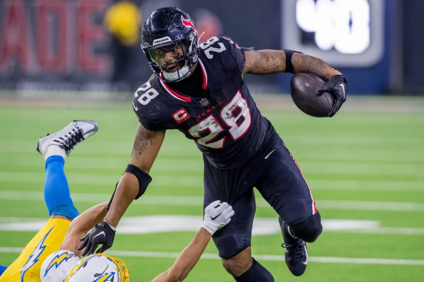 NFL-Star Joe Mixon, in einer schwarzen Uniform mit der Nummer 28, überholt gekonnt einen Gegner in einer weiß-gelben Uniform. Er rennt mit dem Ball über das leuchtend grüne Feld, weicht Tackles mühelos aus und demonstriert so die elektrisierende Intensität des Spiels. Diese Beschreibung wurde mit der FootballR KI automatisch generiert.