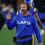 Sean McVay mit einem blauen LAFD-Sweatshirt und einem Headset steht auf einem Footballfeld, möglicherweise der Heimat der Los Angeles Rams. Sie gestikuliert mit der rechten Hand, während im Hintergrund andere Personen in Sportkleidung zu sehen sind. Diese Beschreibung wurde mit der FootballR KI automatisch generiert.