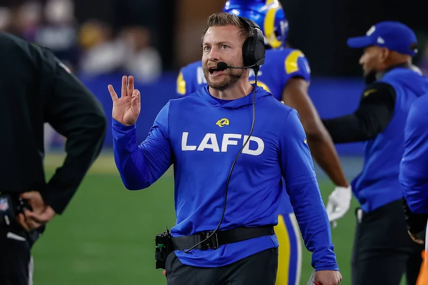 Sean McVay mit einem blauen LAFD-Sweatshirt und einem Headset steht auf einem Footballfeld, möglicherweise der Heimat der Los Angeles Rams. Sie gestikuliert mit der rechten Hand, während im Hintergrund andere Personen in Sportkleidung zu sehen sind. Diese Beschreibung wurde mit der FootballR KI automatisch generiert.