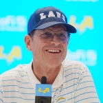 Eine Person mit einer blauen Kappe mit dem Schriftzug „LA“, einer Brille und einem weiß gestreiften Poloshirt steht an einem Podium mit einem Mikrofon, auf dem ein Logo zu sehen ist. Der Hintergrund ist hellblau mit verblassten „SoFi“- und „LA“-Logos. Jim Harbaugh lächelt während einer Pressekonferenz. Diese Beschreibung wurde mit der FootballR KI automatisch generiert.
