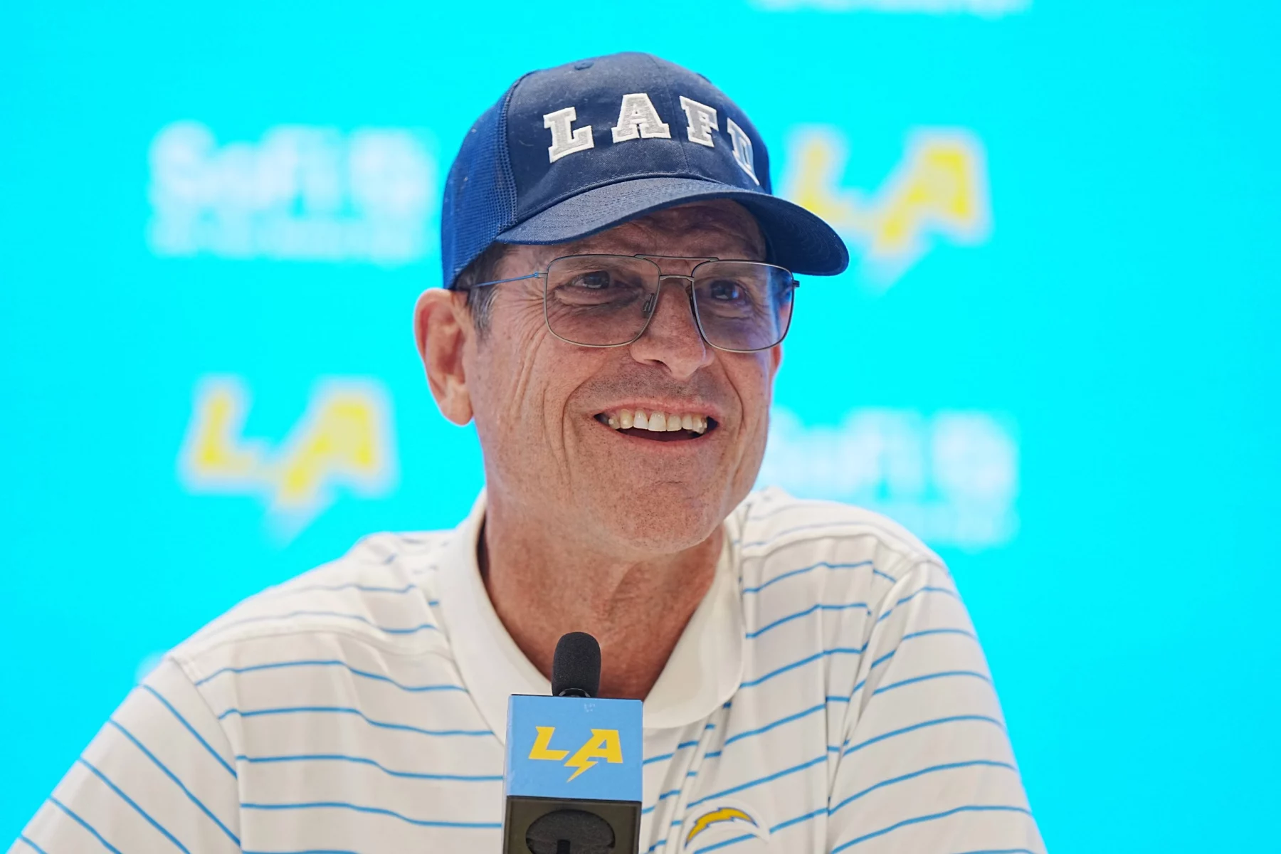 Eine Person mit einer blauen Kappe mit dem Schriftzug „LA“, einer Brille und einem weiß gestreiften Poloshirt steht an einem Podium mit einem Mikrofon, auf dem ein Logo zu sehen ist. Der Hintergrund ist hellblau mit verblassten „SoFi“- und „LA“-Logos. Jim Harbaugh lächelt während einer Pressekonferenz. Diese Beschreibung wurde mit der FootballR KI automatisch generiert.