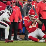 Replay Assists für Quarterback Slides - Ein NFL-Quarterback in rot-weißer Uniform kniet auf dem Feld und blickt auf sein Handgelenk. Ein gegnerischer Spieler in schwarz-weißer Uniform nähert sich, während Trainer und Spieler – einige in roten Jacken – an der Seitenlinie stehen. Das Spiel findet unter dem Nachthimmel statt und wird gespannt verfolgt, um Wiederholungsvorlagen zu erhalten. Diese Beschreibung wurde mit der FootballR KI automatisch generiert.