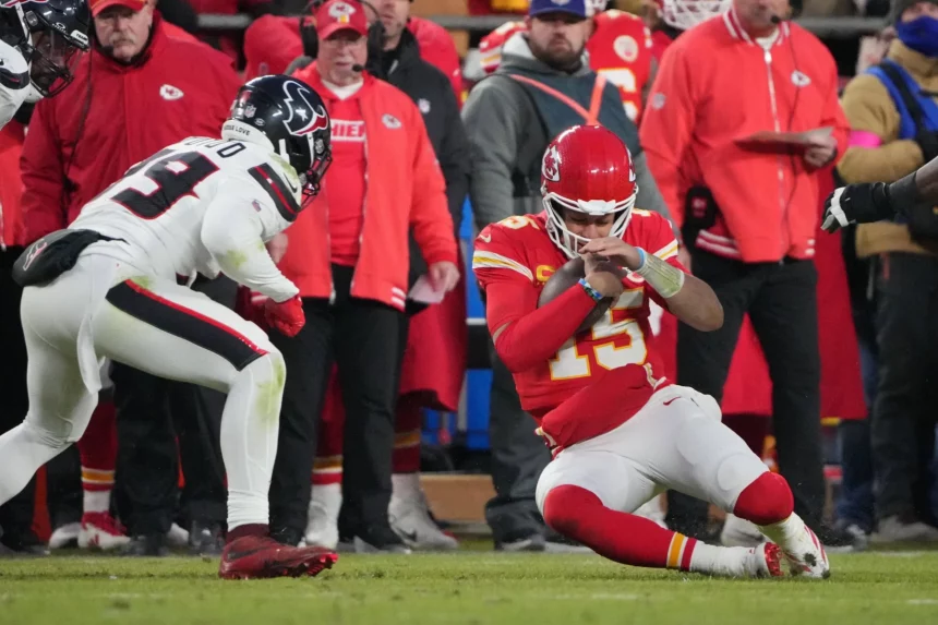 Replay Assists für Quarterback Slides - Ein NFL-Quarterback in rot-weißer Uniform kniet auf dem Feld und blickt auf sein Handgelenk. Ein gegnerischer Spieler in schwarz-weißer Uniform nähert sich, während Trainer und Spieler – einige in roten Jacken – an der Seitenlinie stehen. Das Spiel findet unter dem Nachthimmel statt und wird gespannt verfolgt, um Wiederholungsvorlagen zu erhalten. Diese Beschreibung wurde mit der FootballR KI automatisch generiert.