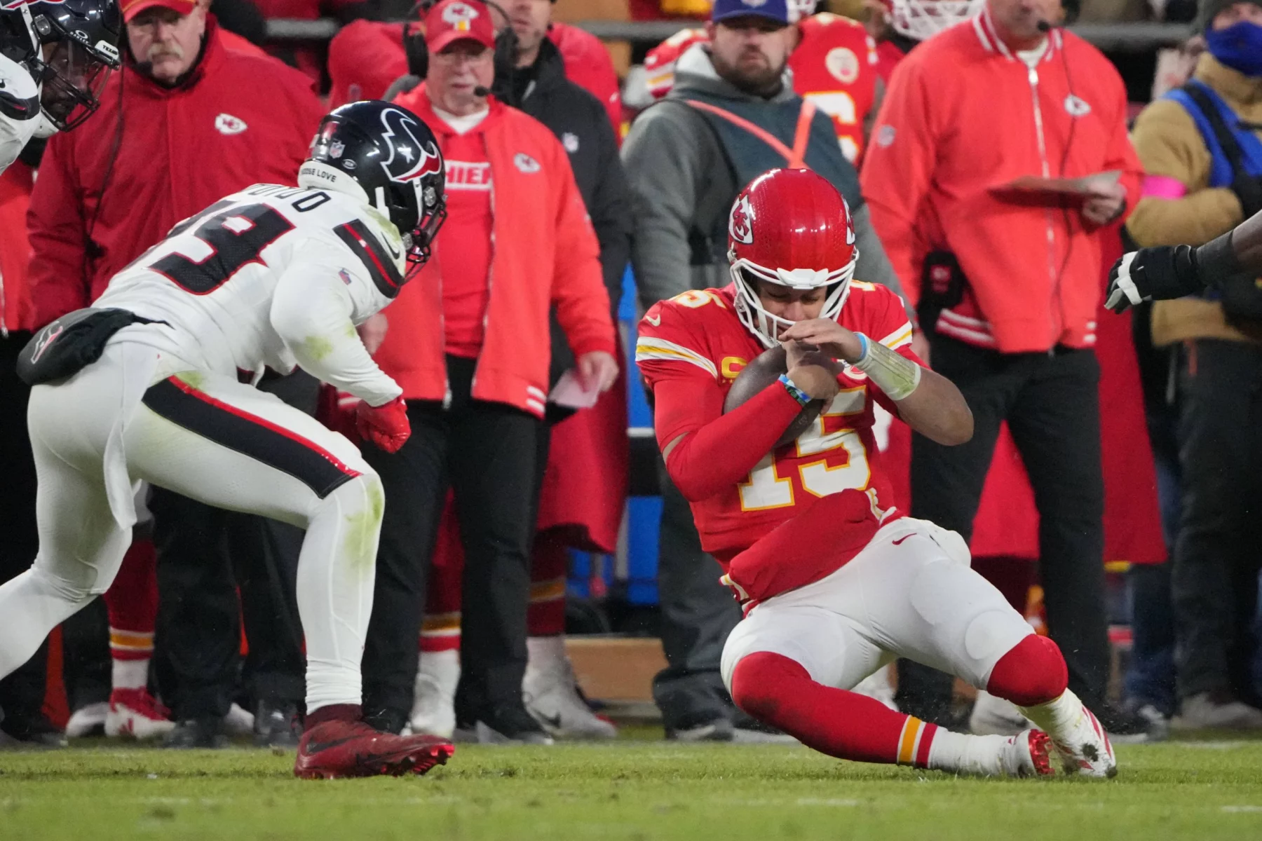 Replay Assists für Quarterback Slides - Ein NFL-Quarterback in rot-weißer Uniform kniet auf dem Feld und blickt auf sein Handgelenk. Ein gegnerischer Spieler in schwarz-weißer Uniform nähert sich, während Trainer und Spieler – einige in roten Jacken – an der Seitenlinie stehen. Das Spiel findet unter dem Nachthimmel statt und wird gespannt verfolgt, um Wiederholungsvorlagen zu erhalten. Diese Beschreibung wurde mit der FootballR KI automatisch generiert.