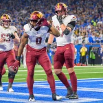 Washington Commanders: Drei Footballspieler in kastanienbraunen Hosen und weißen Trikots jubeln in einer Endzone. Der Spieler in der Mitte posiert, während die anderen beiden zuschauen. Das Stadion ist voller Fans in Blau. Das Spielfeld ist hellblau und weiß markiert. Diese Beschreibung wurde mit der FootballR KI automatisch generiert.