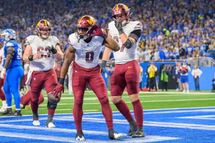 Washington Commanders: Drei Footballspieler in kastanienbraunen Hosen und weißen Trikots jubeln in einer Endzone. Der Spieler in der Mitte posiert, während die anderen beiden zuschauen. Das Stadion ist voller Fans in Blau. Das Spielfeld ist hellblau und weiß markiert. Diese Beschreibung wurde mit der FootballR KI automatisch generiert.