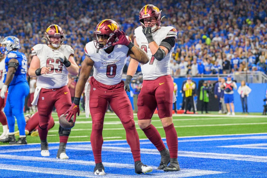 Washington Commanders: Drei Footballspieler in kastanienbraunen Hosen und weißen Trikots jubeln in einer Endzone. Der Spieler in der Mitte posiert, während die anderen beiden zuschauen. Das Stadion ist voller Fans in Blau. Das Spielfeld ist hellblau und weiß markiert. Diese Beschreibung wurde mit der FootballR KI automatisch generiert.