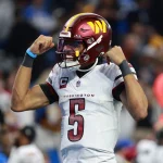 Rookie-Quarterback Jayden Daniels, trägt eine Uniform der Washington Commanders mit der Nummer 5 und feiert. Er trägt ein weißes Trikot, einen burgunderfarbenen Helm und spannt beide Arme an. Der Hintergrund ist verschwommen, einige Personen sind zu sehen und eine rote Seitenlinienmarkierung ist zu sehen. Diese Beschreibung wurde mit der FootballR KI automatisch generiert.